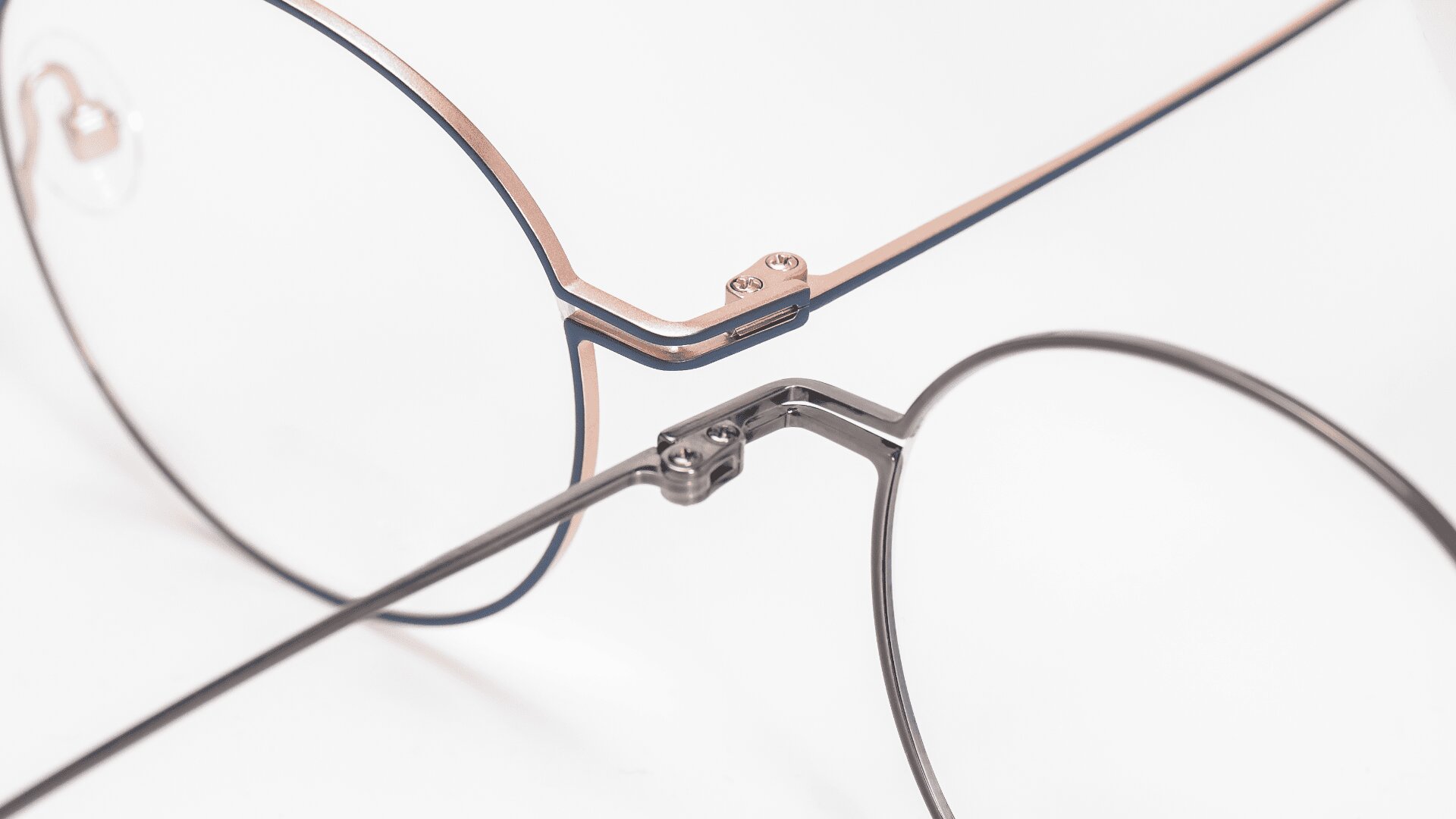 Nahaufnahme von zwei Brillen, die sich auf weißem Hintergrund überlappen, wobei die Metallrahmen und Scharniere im Detail hervorgehoben werden. Eine Brille hat einen dünnen, silbernen Rahmen, die andere hat einen dickeren, blau-goldenen Rahmen.