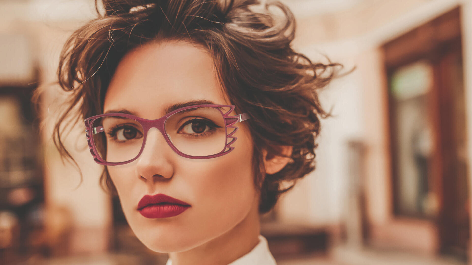 Eine Person mit kurzem, welligem braunem Haar, kräftigem roten Lippenstift und stylischer lila Brille mit einzigartigem Flügelrahmen, blickt in einer leicht verschwommenen Innenkulisse selbstbewusst in die Kamera.