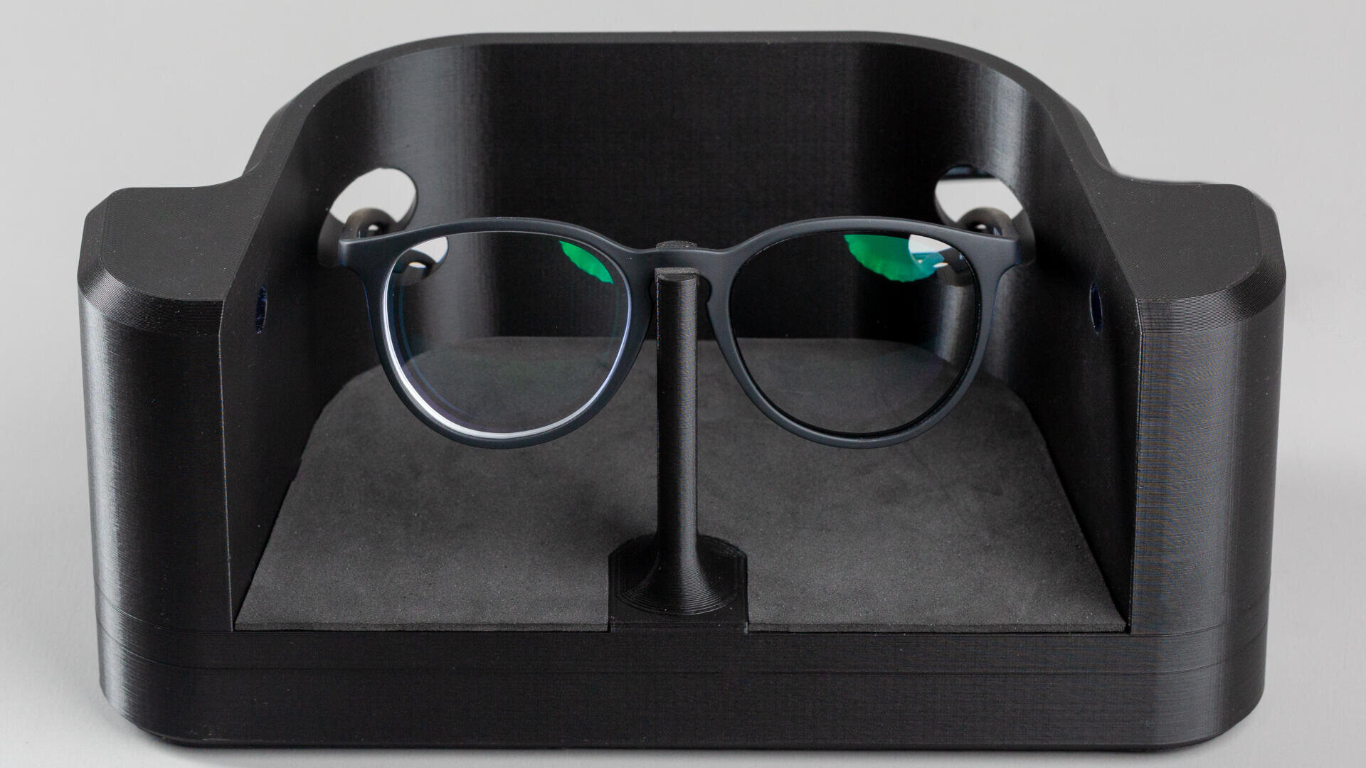 Eine schwarze Brille, aufrecht auf einem Ständer in einem schwarzen, rechteckigen Halter mit abgerundeten Kanten und einer weichen, grauen Innenbasis positioniert.