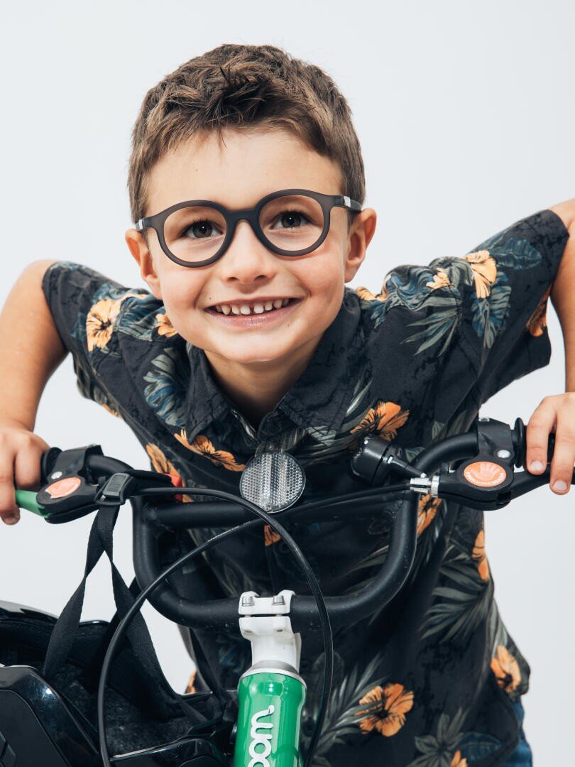 Ein lächelnder kleiner Junge mit Brille und geblümtem Hemd hält den Lenker eines Fahrrads und blickt vor einem schlichten weißen Hintergrund direkt in die Kamera.