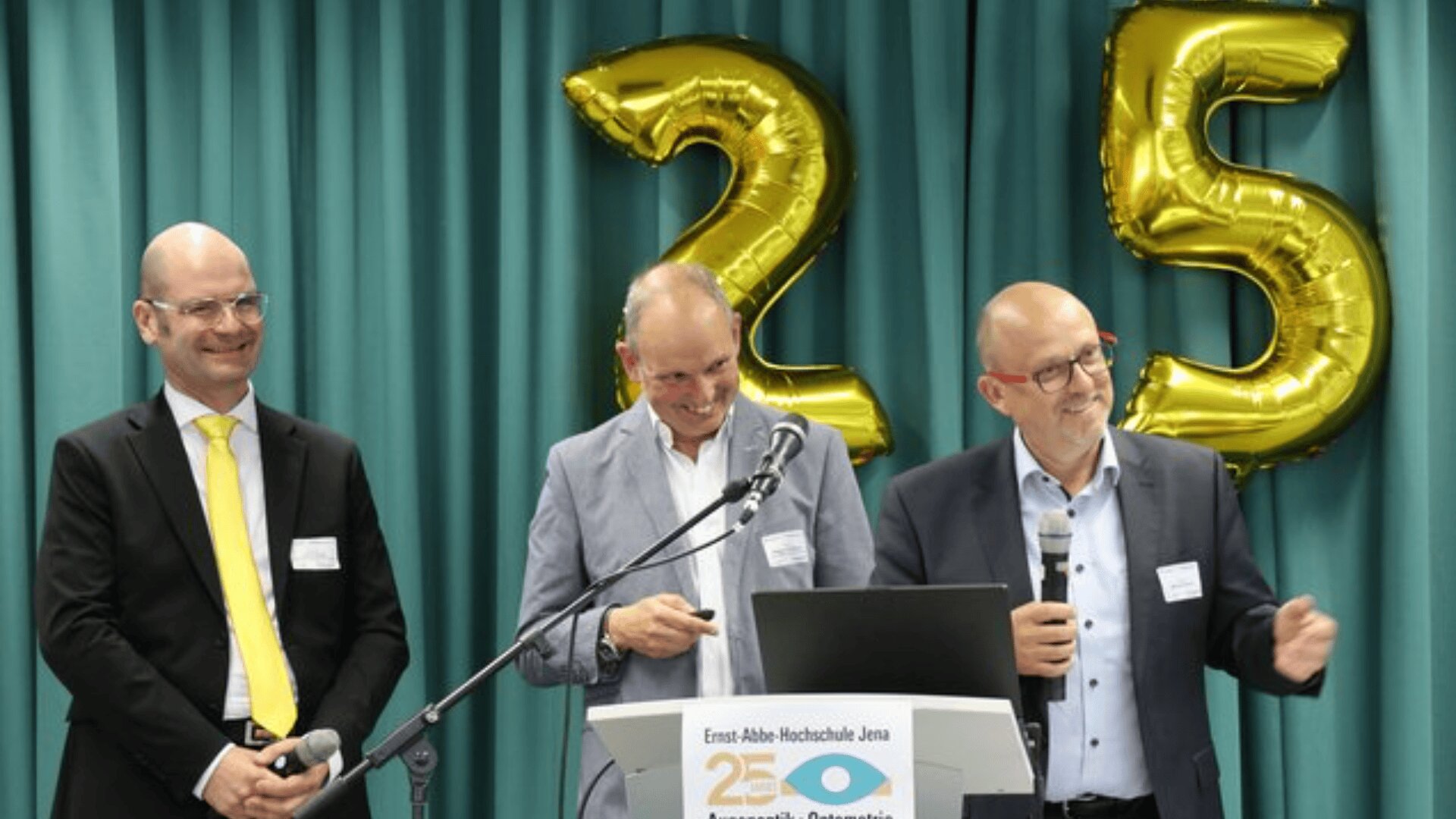 Drei Männer in Anzügen stehen lächelnd an einem Podium mit Mikrofonen, vor großen goldenen Luftballons in Form der Zahl 25. Auf einem Schild am Podium steht "Ernst Abbe Hochschule Jena". Im Hintergrund hängen grüne Vorhänge.
