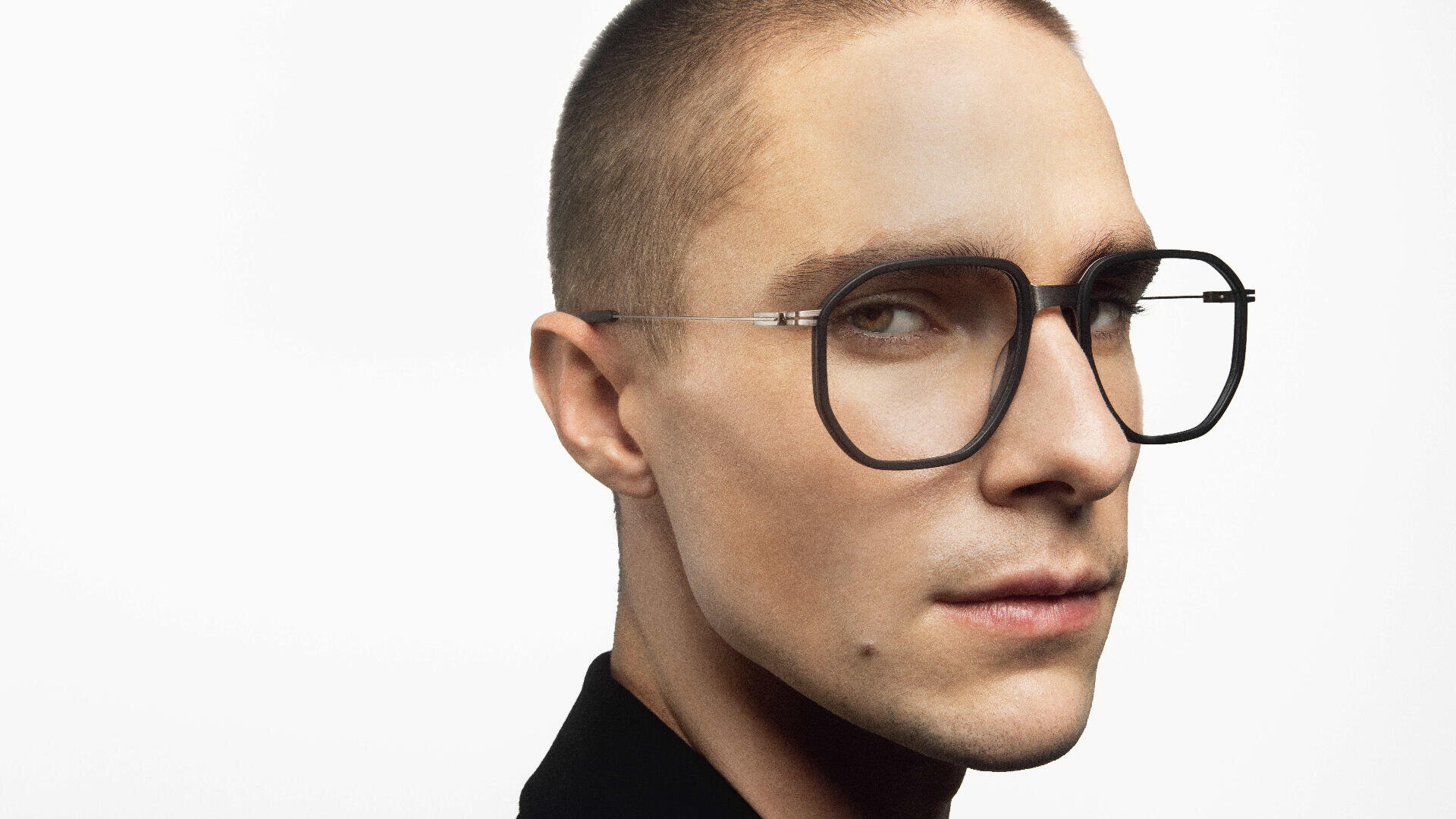 A person with short hair and light skin wears large, round, black-rimmed eyeglasses and a black top, looking confidently at the camera against a plain white background.