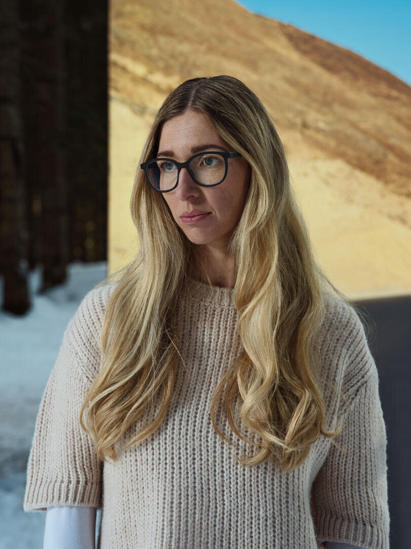 Eine Frau mit langen blonden Haaren und Brille steht im Freien und trägt einen beigen Strickpullover. Hinter ihr ist ein kontrastreicher Hintergrund mit einem sonnenbeschienenen Hügel und dunklen, schneebedeckten Bäumen zu sehen.