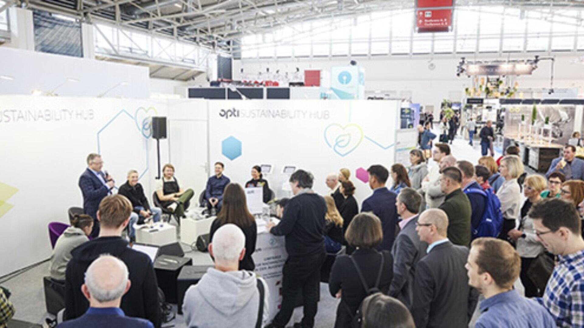 A group of people attends a presentation at an indoor exhibition. The presenter stands next to a panel, while the audience listens attentively in front of booths labeled “Sustainability Hub.”.