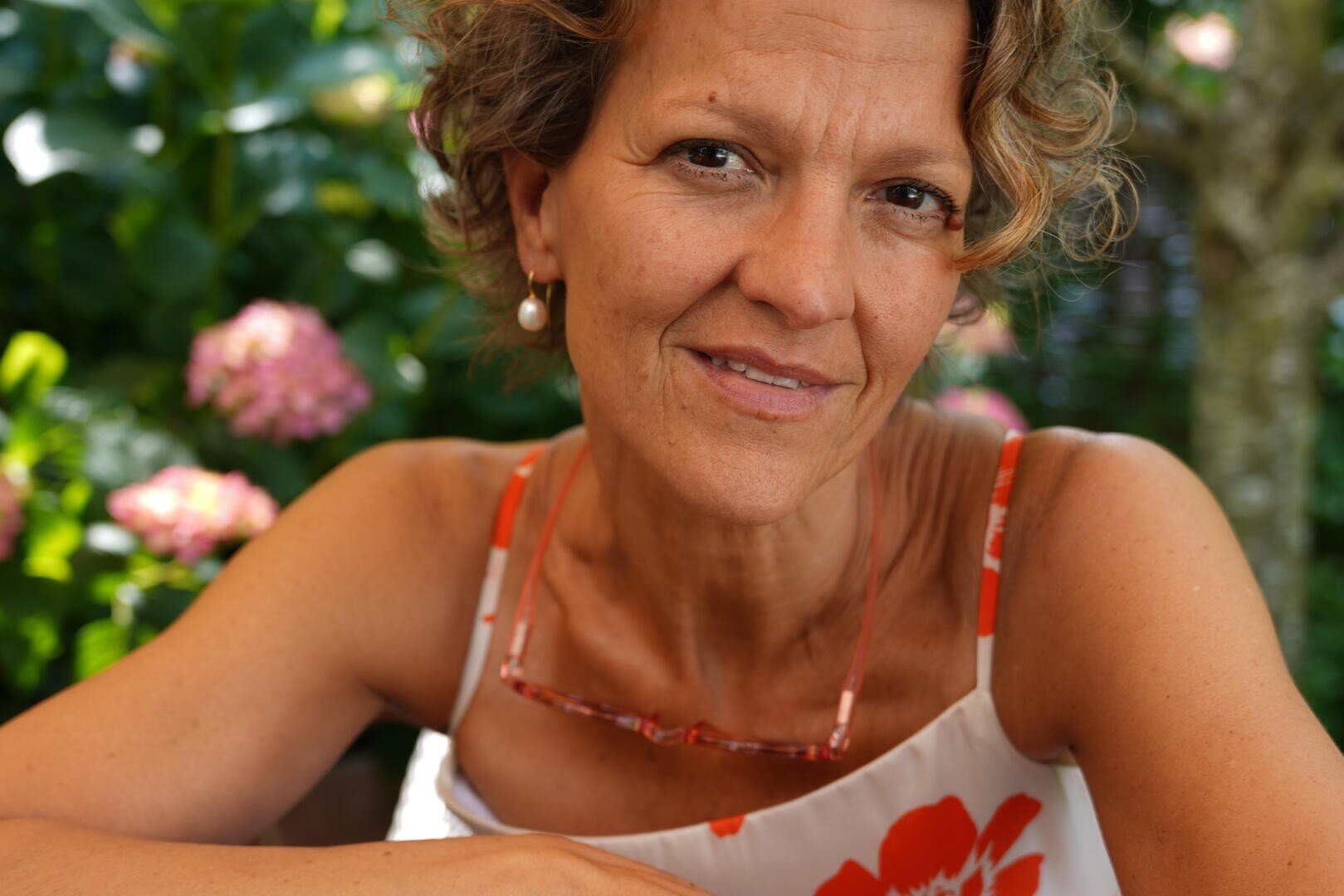 A middle-aged woman with curly hair, wearing a flowered dress and pearl earrings, smiles gently as she sits outside in front of blooming pink flowers and green foliage.