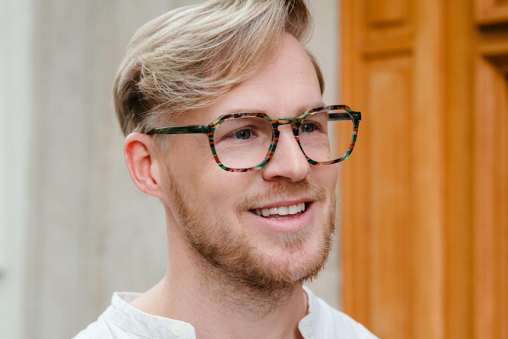 Ein lächelnder Mann mit hellblondem Haar und kurzem Bart trägt eine große Schildpattbrille und ein weißes Hemd und steht vor einer Holztür und einer hellen Wand.