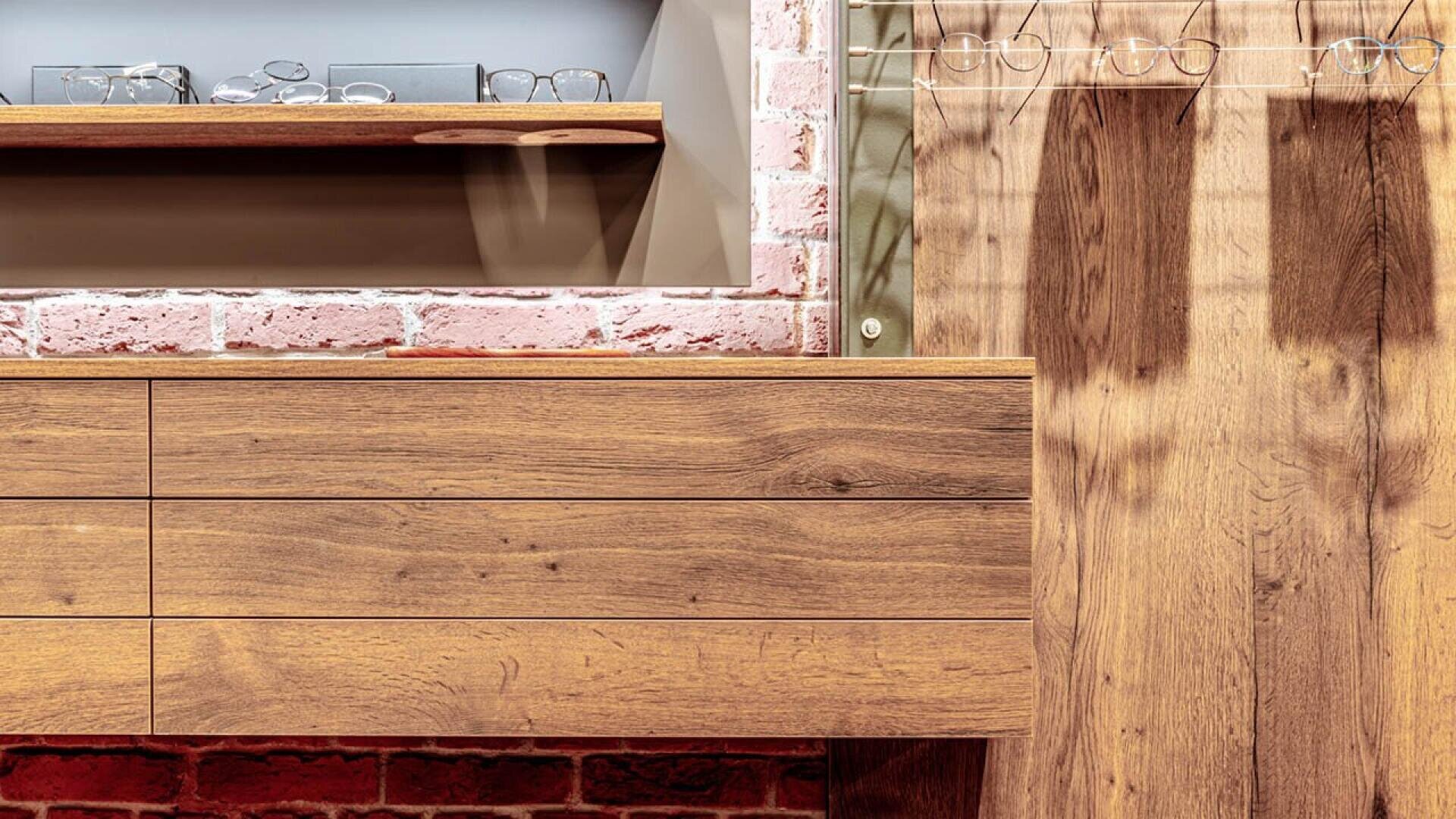 A modern interior with wooden drawers and cabinet, a shelf holding eyeglasses, a coat rack with more eyeglasses, and a brick wall background. Warm tones dominate the scene.