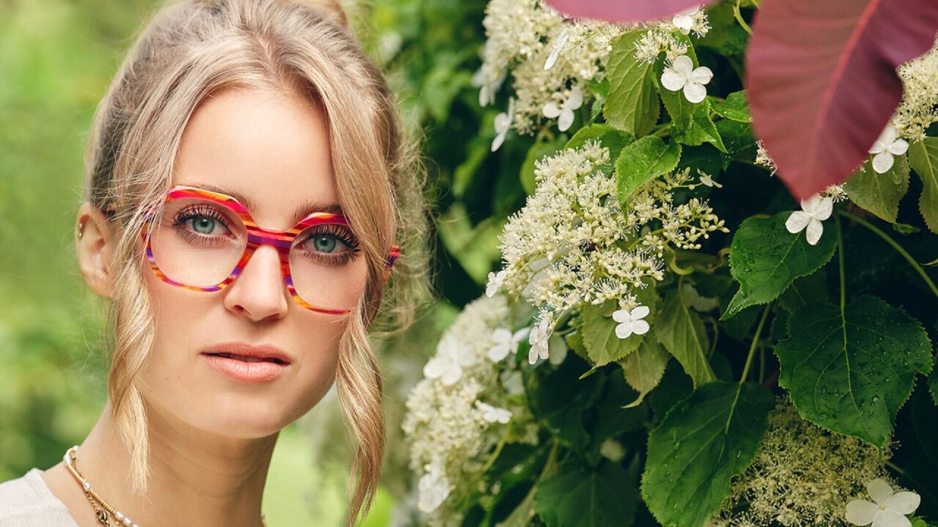 A woman with blonde hair in a loose bun, wearing colorful red and yellow glasses, stands next to green foliage and white flowers, looking directly at the camera.