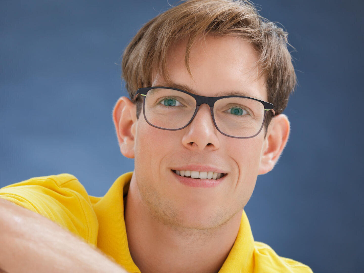 Ein lächelnder Mann mit hellbraunem Haar und Brille, der ein gelbes Poloshirt trägt, posiert vor einem blauen Hintergrund.