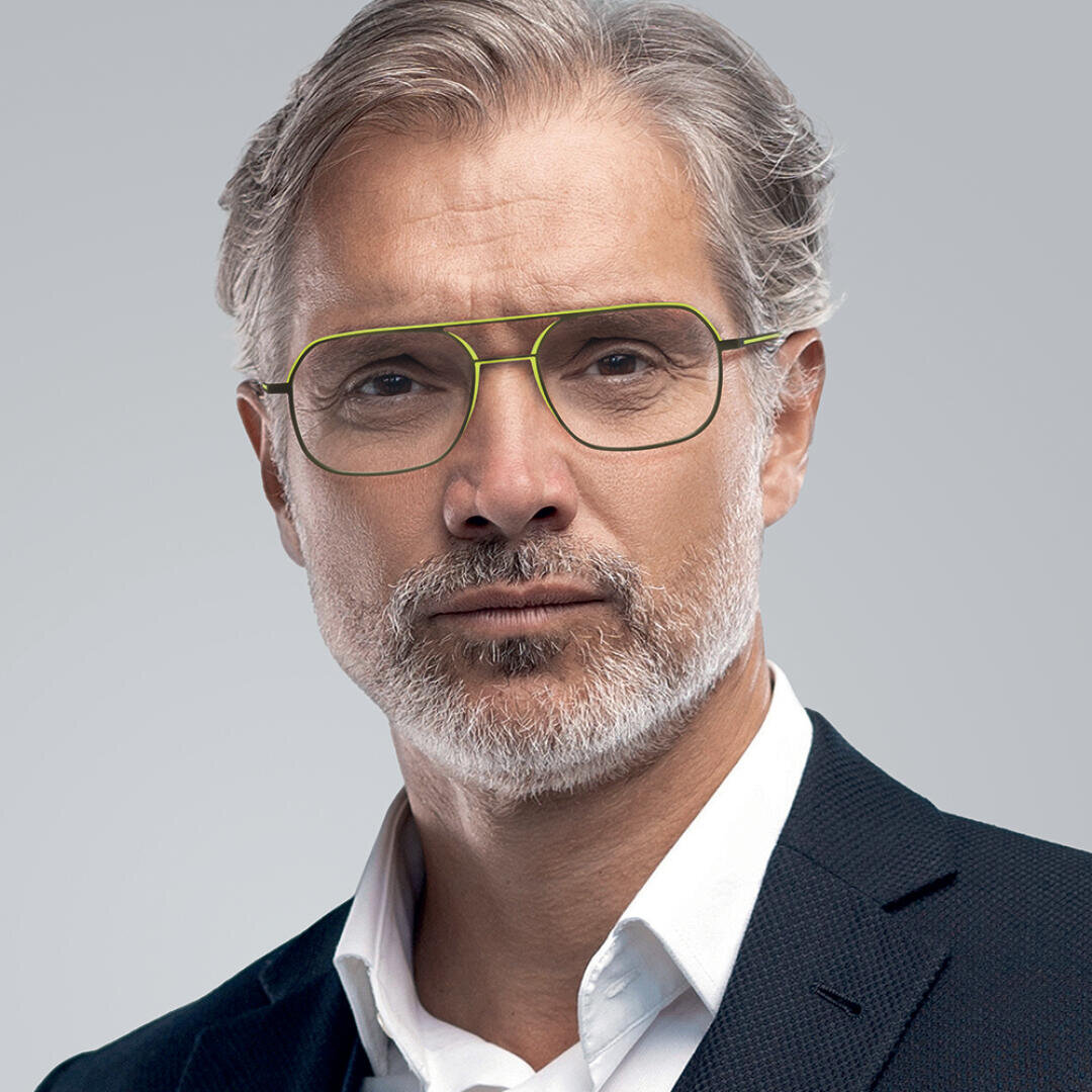 A middle-aged man with gray hair and a short beard wears yellow-rimmed glasses and a dark suit jacket over a white shirt, posing against a plain gray background.