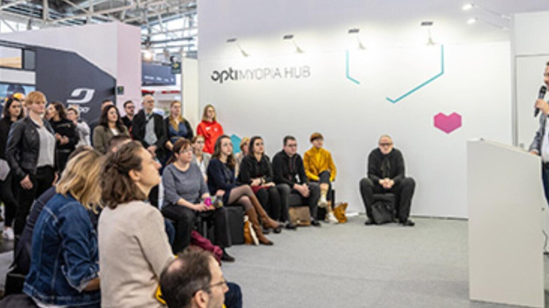 A group of people sit and stand attentively in an indoor event space, listening to a speaker at a podium. The backdrop reads OPTI MYOPIA HUB with geometric designs on the wall.