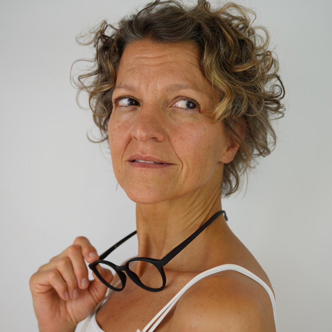 A middle-aged woman with curly short hair holds a pair of black glasses in one hand. She wears a white tank top and looks slightly to the side with a thoughtful expression against a plain light-colored background.