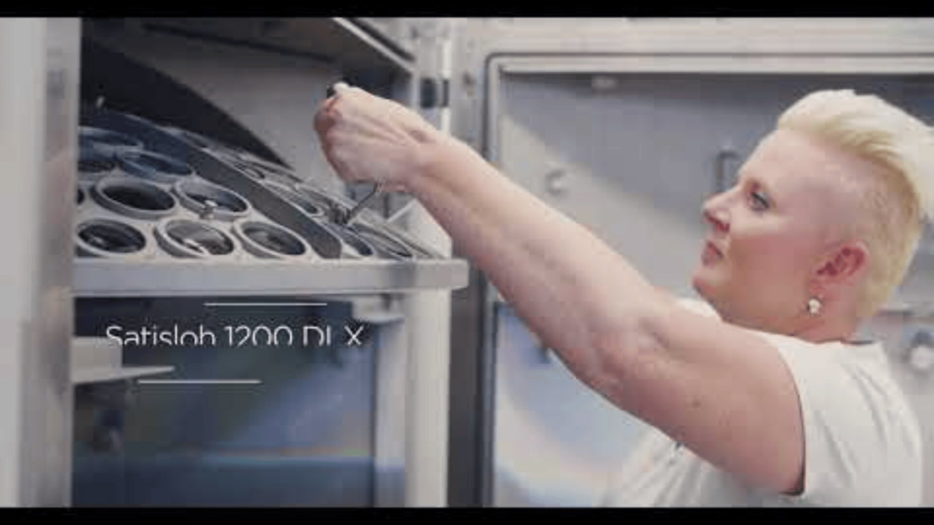Eine Person mit kurzen blonden Haaren stellt Geräte in einer großen Industriemaschine mit der Aufschrift „Satisloh 1200 DL X“ ein.