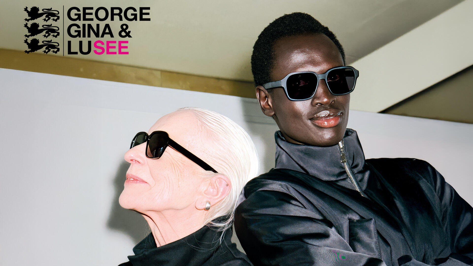 Two stylish people with dark sunglasses and black jackets pose confidently in front of a white background. The George Gina & Lucy logo appears in the top left corner.