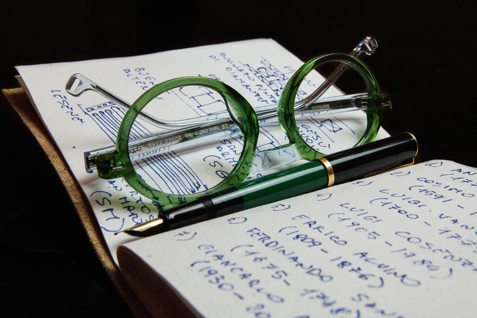 A pair of green-rimmed glasses and a green fountain pen lie on top of an open notebook filled with handwritten notes in blue ink.