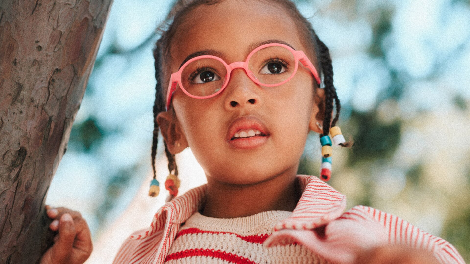 Ein kleines Kind mit geflochtenen Haaren und bunten Perlen trägt eine rosafarbene Brille und einen gestreiften Pullover und blickt mit neugierigem Blick nach oben. Bäume und Sonnenlicht bilden einen weichen, verschwommenen Hintergrund.