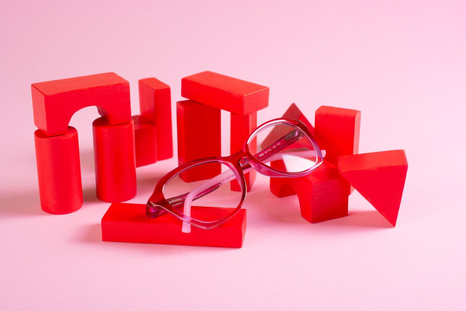 A pair of red eyeglasses rests on red geometric wooden blocks arranged on a pink surface with a pink background.