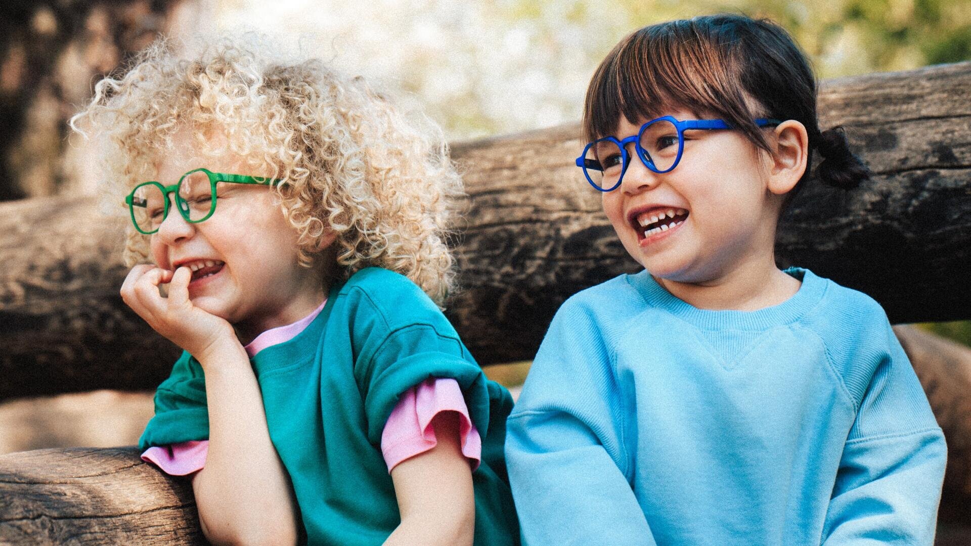Two small children with colorful glasses and a smile are sitting outside on a wooden fence. One child has curly blond hair and a green shirt, the other has straight brown hair and is wearing a light blue sweatshirt.