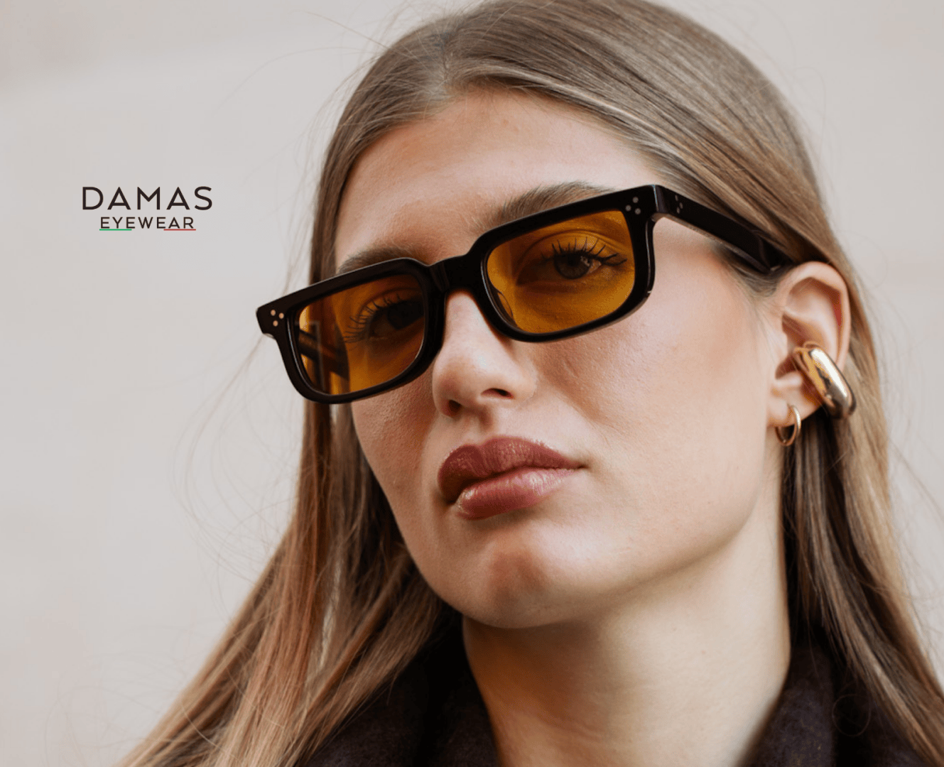 A woman wearing rectangular, black-framed yellow tinted sunglasses and a chunky gold ear cuff. The image includes the DAMAS EYEWEAR logo on a neutral background.