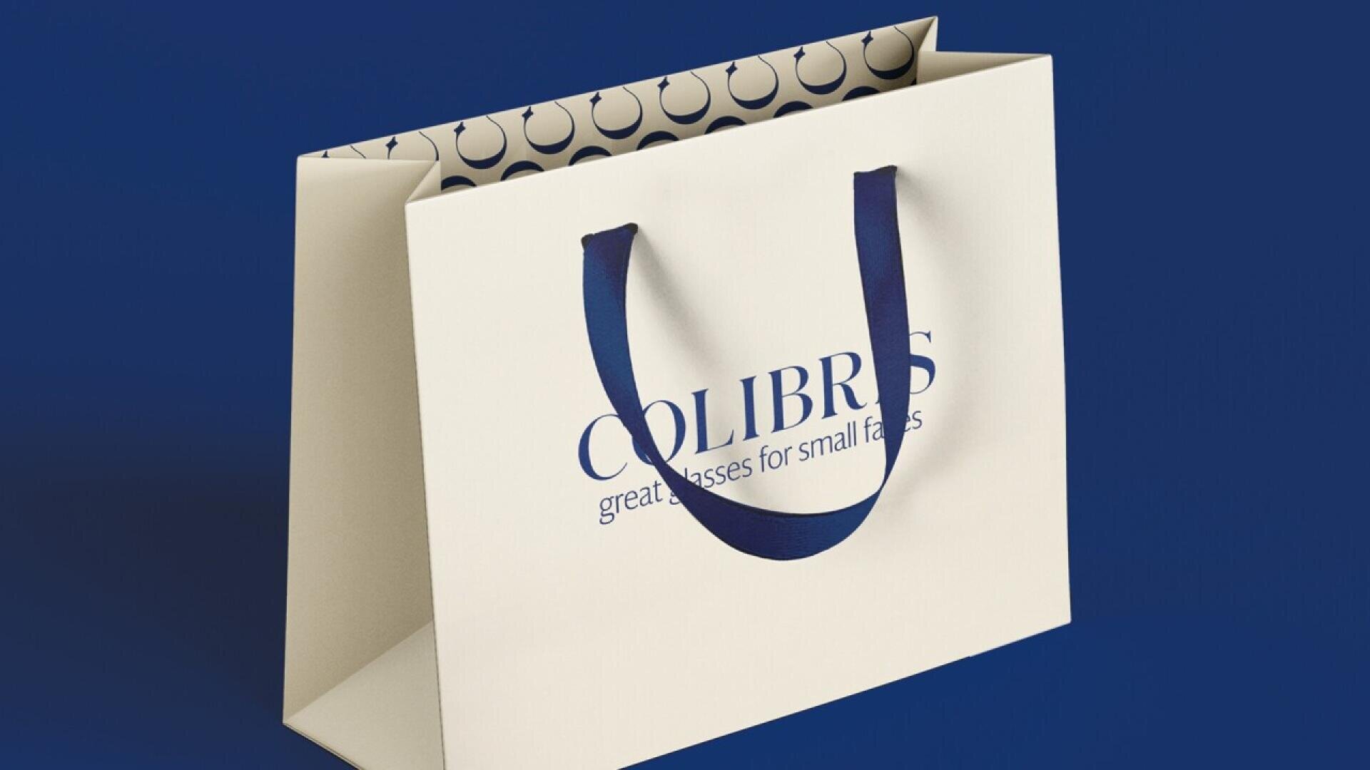 A cream-colored shopping bag with dark blue handles sits on a blue background. The bag features the text “COLIBRIS great glasses for small faces” and has a patterned interior with circular designs.