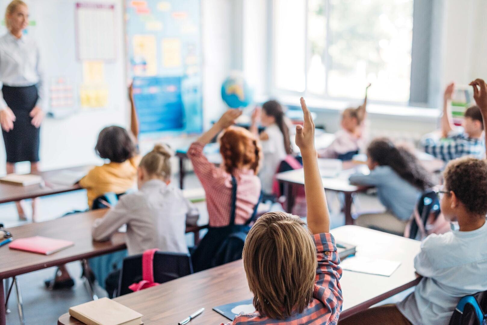 Ein Klassenzimmer mit jungen Schülern, die an den Tischen sitzen und ihre Hände heben, um eine Frage zu beantworten. Eine Lehrerin steht an der Stirnseite des Raumes, und durch große Fenster im Hintergrund fällt Sonnenlicht.