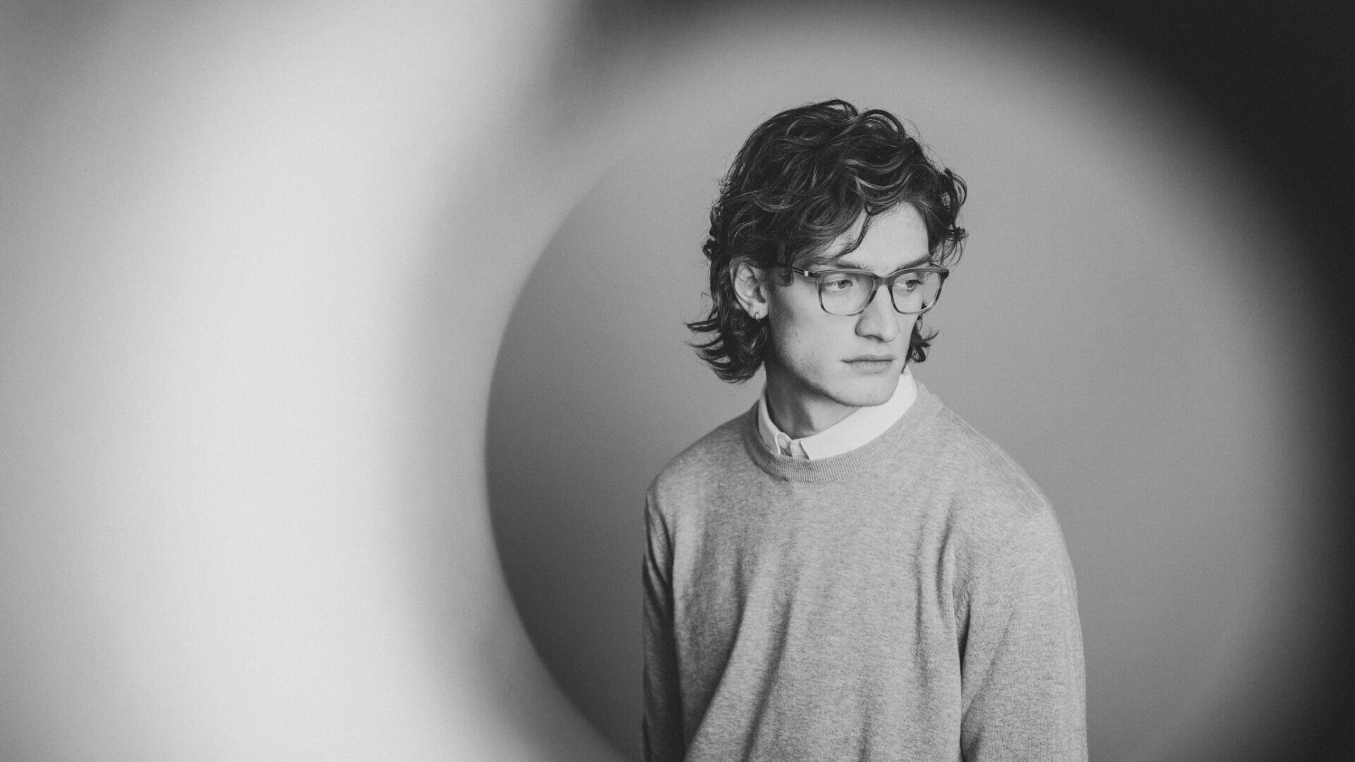 Eine junge Person mit welligem Haar und Brille, die einen hellen Pullover über einem Hemd mit Kragen trägt, blickt nachdenklich zur Seite. Das Foto ist schwarz-weiß mit einem unscharfen kreisförmigen Vignetteneffekt.