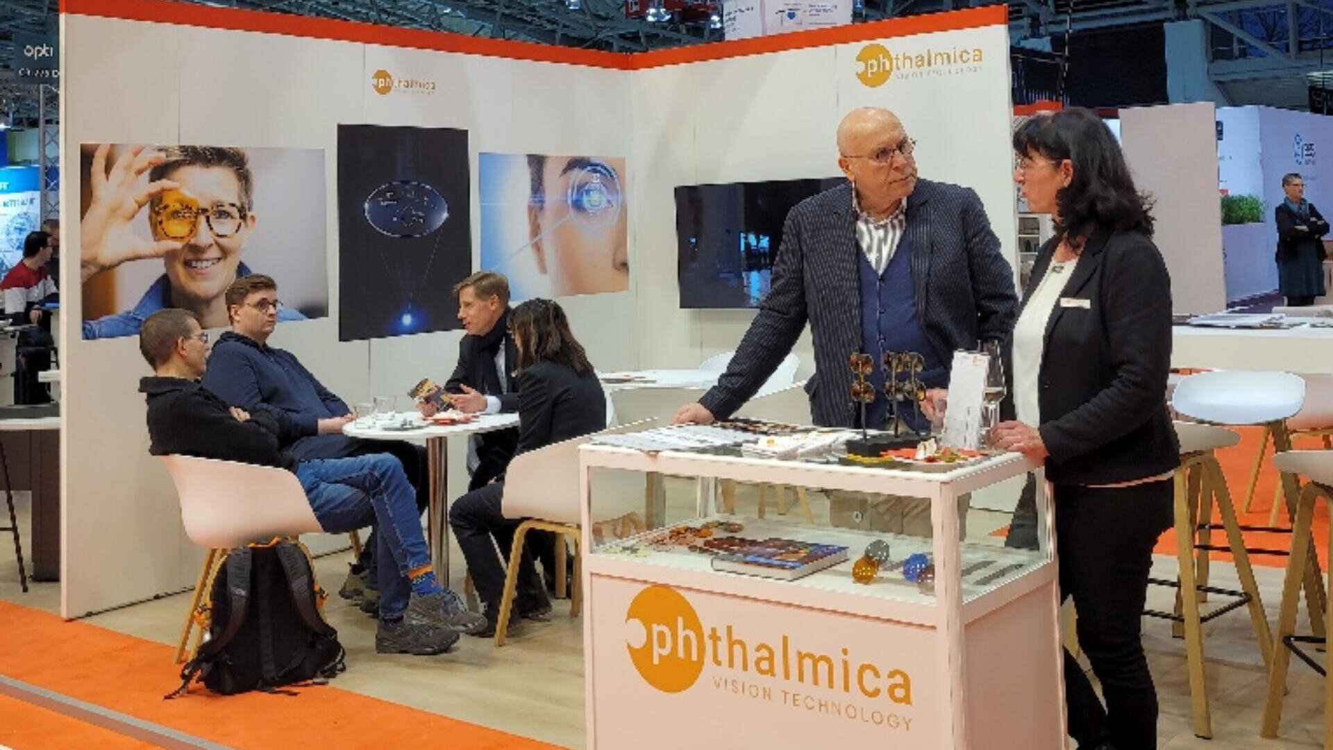 Am Messestand von Ophthalmica Vision Technology sitzen Menschen an Tischen und unterhalten sich. Auf einem Tresen sind Werbedisplays und Produktmuster zu sehen, im Hintergrund sind Markenbanner zu sehen.