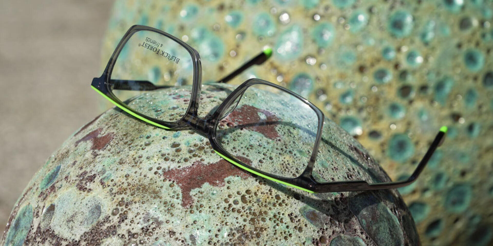 Eine Brille mit schwarzem und grünem Rahmen ruht auf einer grünlichen, verwitterten Steinkugel mit Struktur. Eine weitere ähnliche Kugel erscheint im unscharfen Hintergrund.