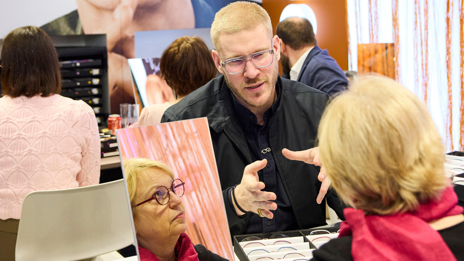 Ein Mann mit Brille gestikuliert, während er mit einer älteren Frau spricht, die ebenfalls eine Brille trägt und in einen Spiegel blickt. Sie sitzen an einem Tisch mit Brillengestellen in einer hell erleuchteten Optikerpraxis.