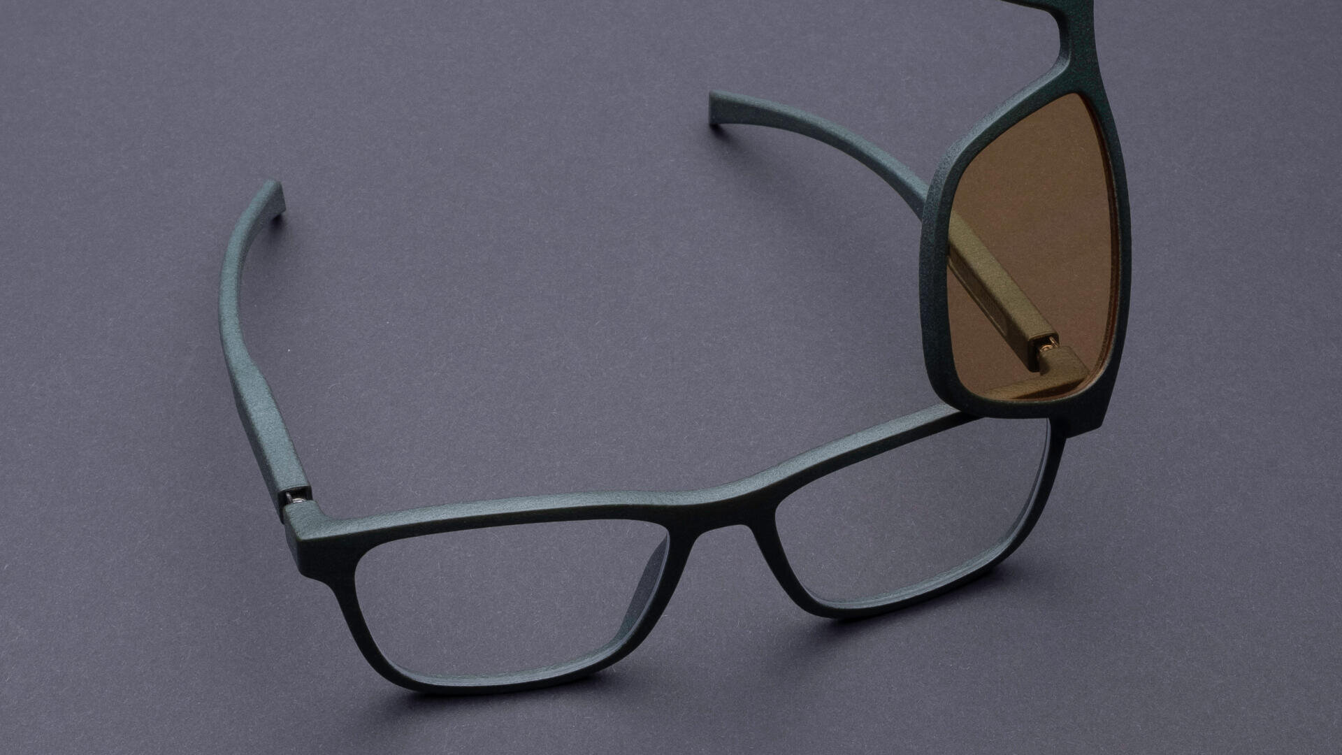 Eine rechteckige Brille mit dunkelgrünem Rahmen, abgebildet mit teilweise angebrachten braun getönten Aufsteckgläsern auf einer dunkelgrauen Oberfläche.
