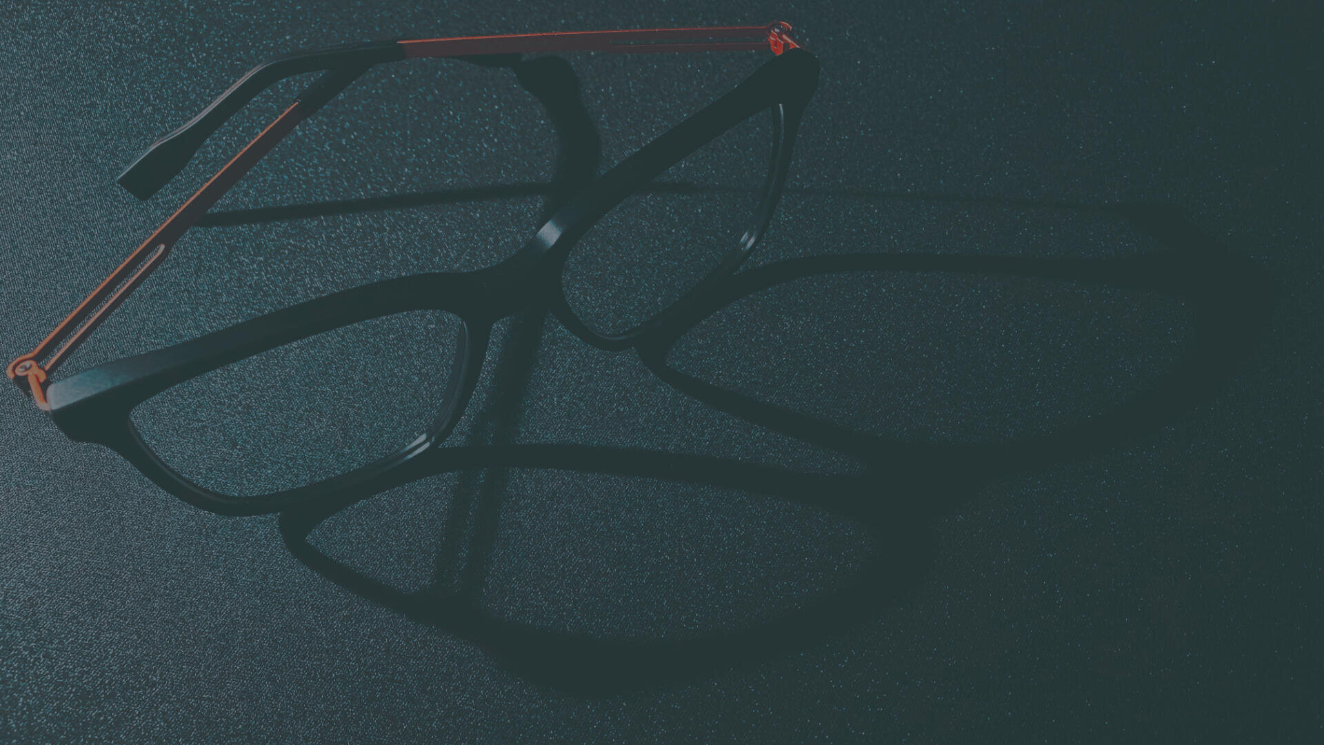 Eine schwarze Brille mit roten Bügeln wirft bei dramatischer Beleuchtung einen markanten Schatten auf eine dunkle, strukturierte Oberfläche.
