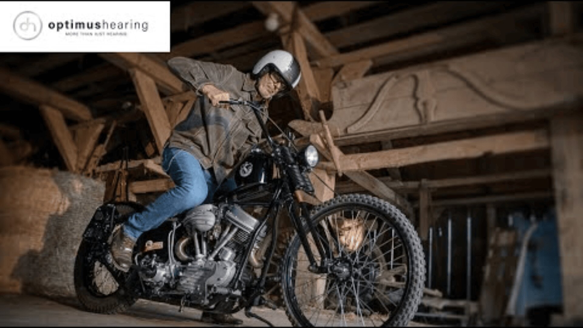 Ein Mann mit Helm und Brille startet in einer rustikalen Holzscheune ein Oldtimer-Motorrad. Das Optimus-Logo erscheint in der oberen linken Ecke.