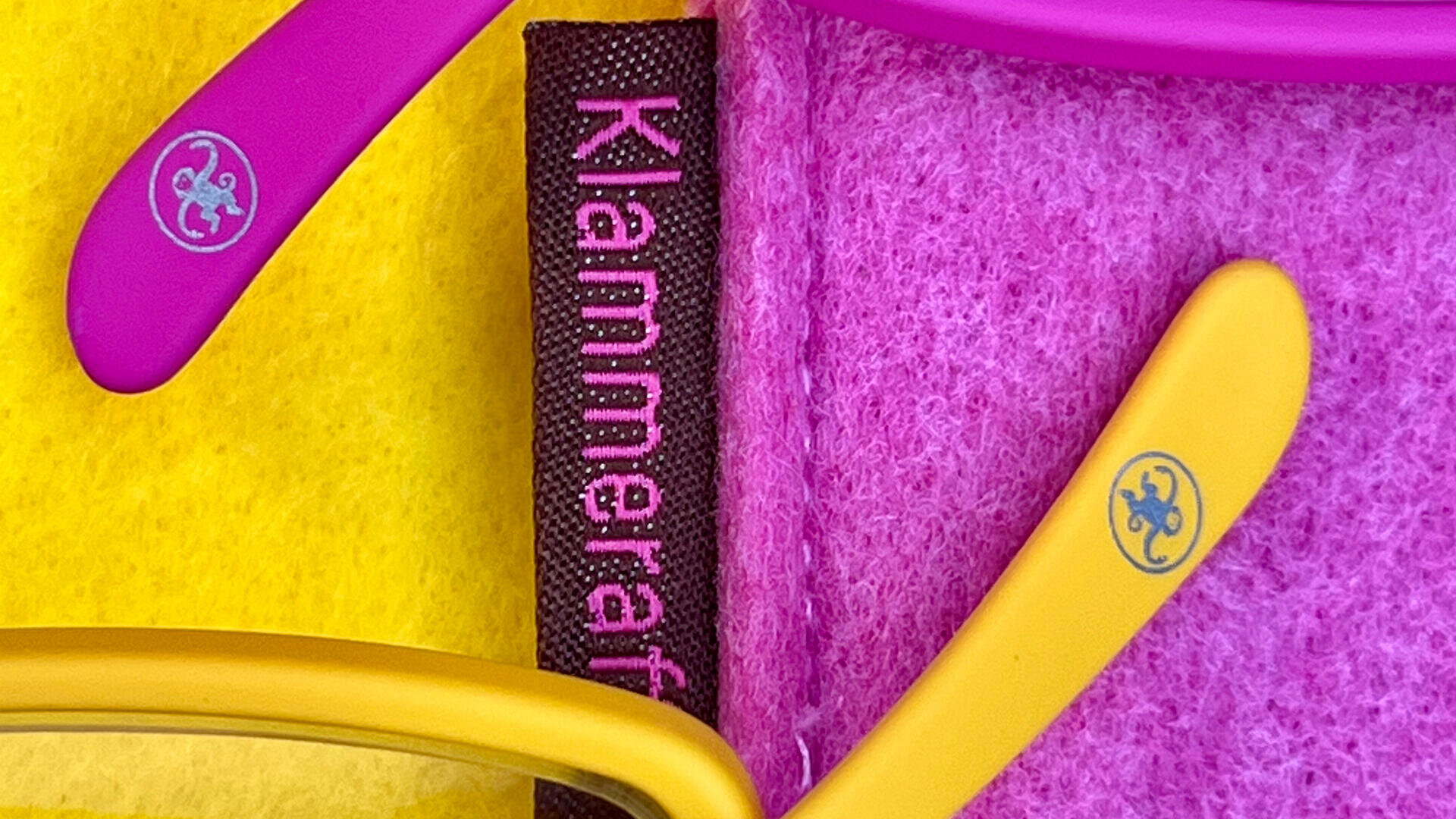 Eine Nahaufnahme von gelben und rosafarbenen Brillenbügeln mit kleinen runden Logos, die auf gelbem und rosafarbenem Filz liegen. Dazwischen befindet sich ein schwarzer Streifen mit dem vertikal in rosafarbener Schrift geschriebenen Wort „Klammeraf“.