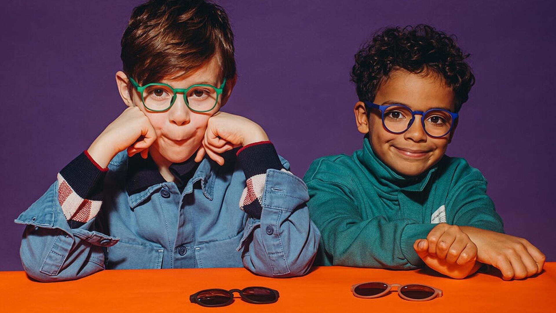 Two young boys wearing colorful glasses sit at an orange table against a purple background. One boy rests his chin on his hands, while the other smiles with his arm outstretched. Two pairs of sunglasses sit on the table.