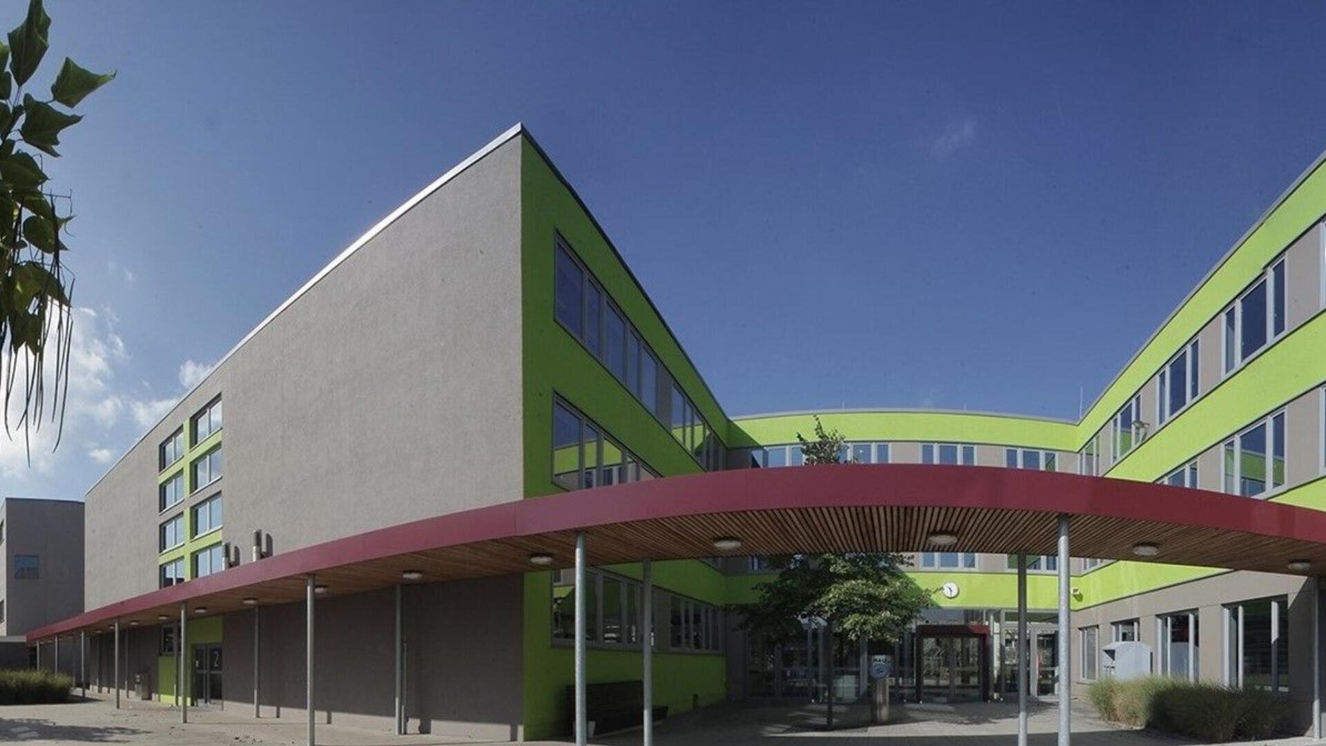 Ein modernes, dreistöckiges Gebäude mit grauen und hellgrünen Wänden, großen Fenstern und einem roten Vordach am Eingang vor einem klaren blauen Himmel.