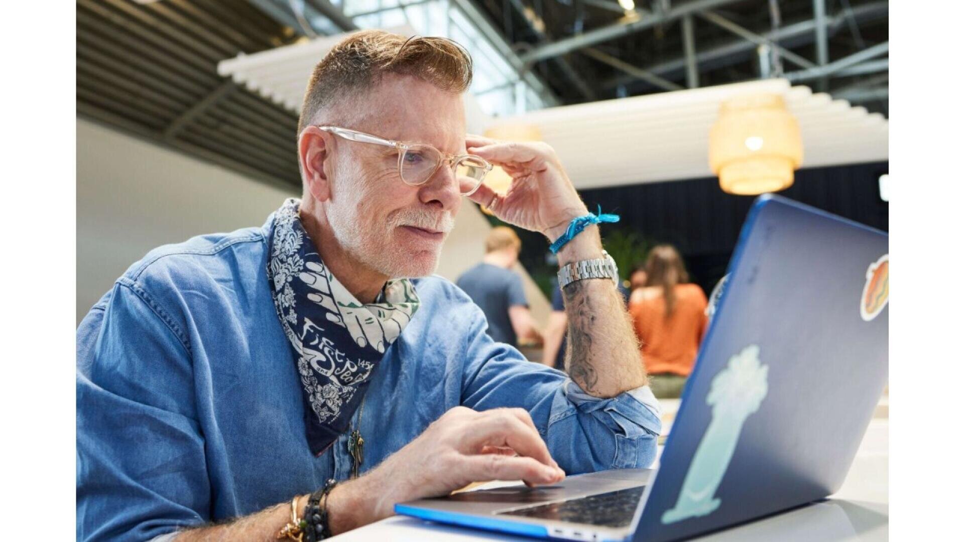 A man wearing glasses and a denim shirt with a patterned scarf sits at a table, focusing on his laptop. People gather in the blurred background of a modern, open space with high ceilings.