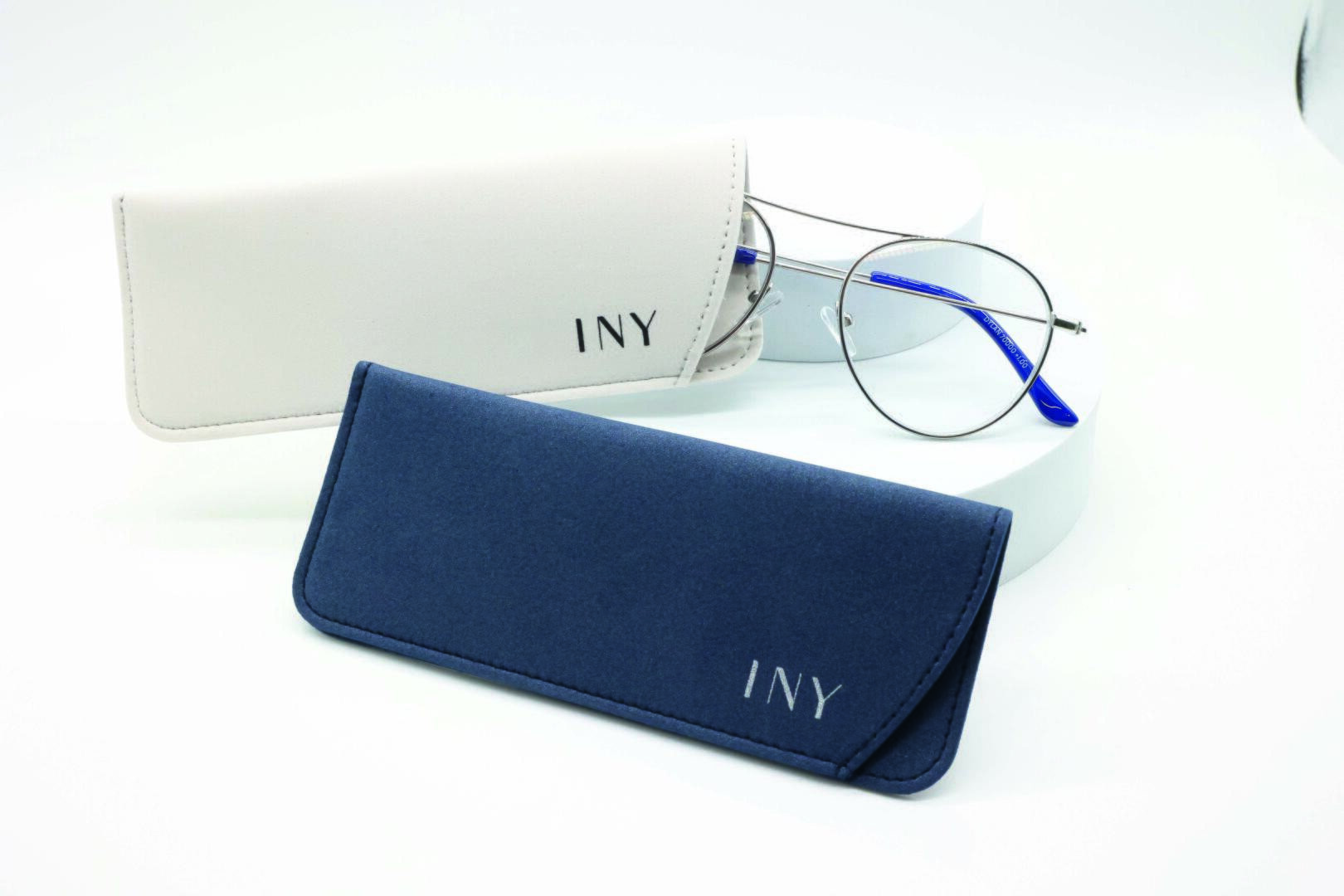 Eine Brille mit silbernem Rand und blauen Bügelenden liegt neben zwei weichen Brillenetuis, eines in Beige und eines in Marineblau, beide mit der Aufschrift „INY“ auf weißem Untergrund.