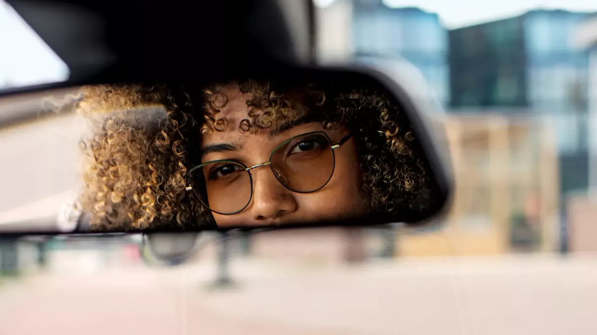 Nahaufnahme einer Person mit lockigem Haar und Brille, die teilweise im Rückspiegel eines Autos sichtbar ist und die Spiegelung vor einem unscharfen Außenhintergrund betrachtet.