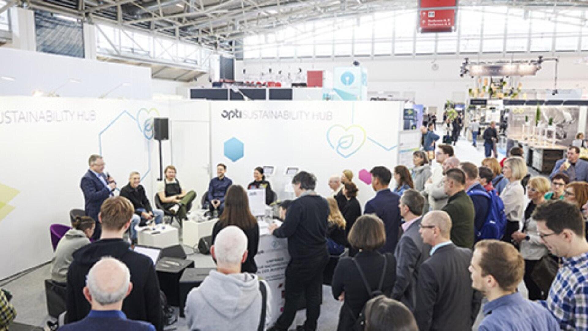A large group of people attend a presentation or panel discussion at a trade show or conference, seated and standing in front of a white booth labeled Sustainability Hub with speakers seated at the front.