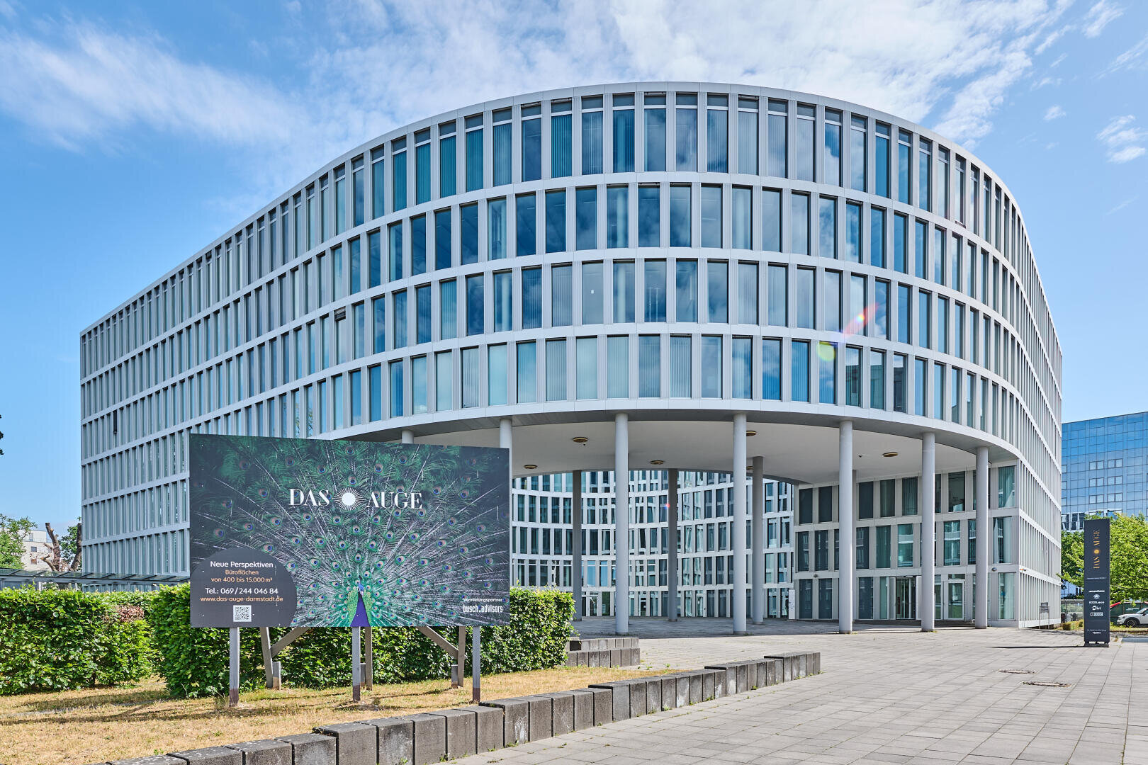 Ein modernes, geschwungenes Bürogebäude aus Glas mit weißer Umrahmung steht hinter einer großen Werbetafel und Grünpflanzen unter einem blauen Himmel mit vereinzelten Wolken.