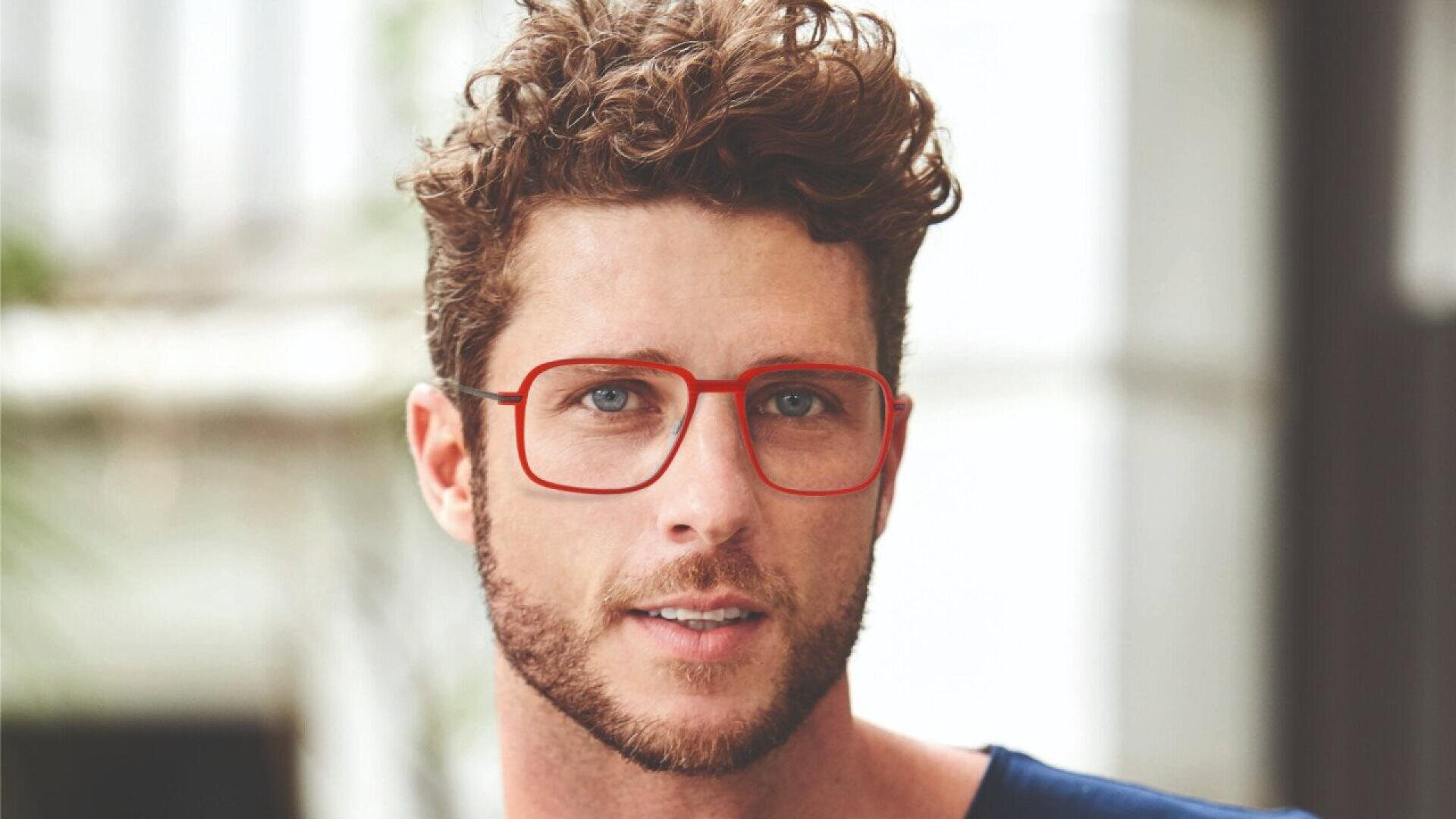 A man with curly brown hair, a trimmed beard, and red glasses looks at the camera. He is wearing a dark blue shirt and is outdoors with a blurred background.