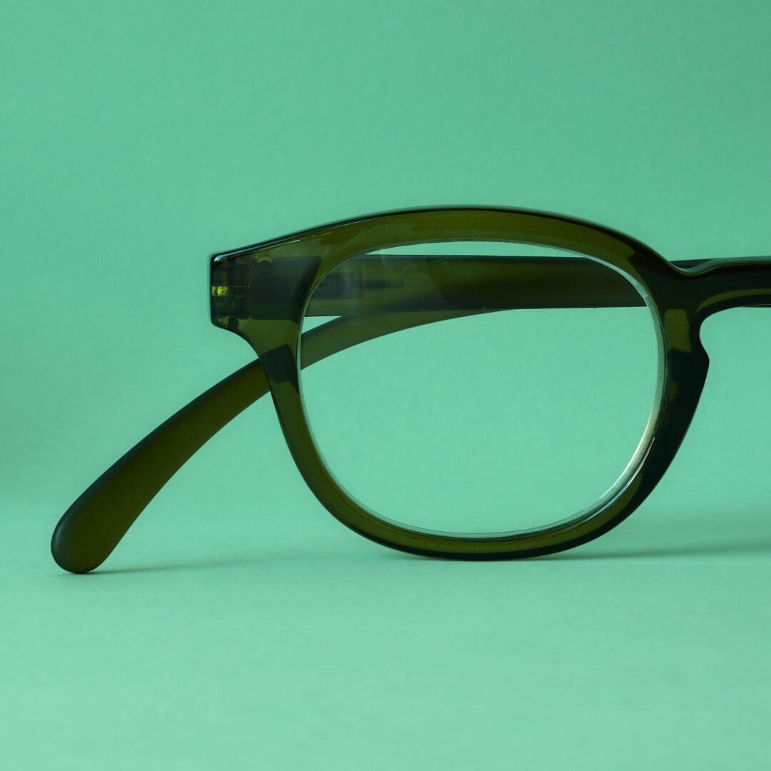 A close-up of dark green glasses with thick frames, set against a matching light green background. Only the right side of the glasses is visible.
