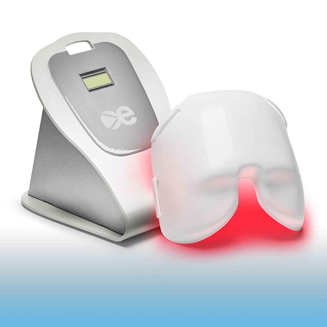Eine weiße und silberne LED-Lichttherapie-Gesichtsmaske sitzt auf ihrem Ladeständer und strahlt ein rotes Licht aus dem Inneren der Maske ab. Der Ständer hat eine kleine Digitalanzeige und ein Logo auf der Vorderseite.