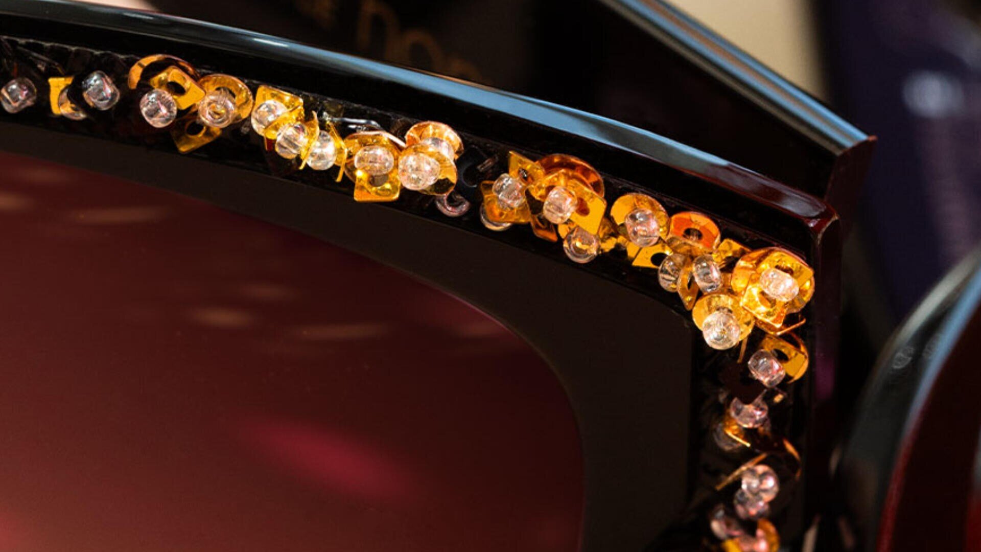 Nahaufnahme einer schwarzen Sonnenbrille, deren oberer Rahmen mit funkelnden gelben, orangefarbenen und klaren Edelsteinen verziert ist, was für ein luxuriöses und stilvolles Aussehen sorgt.