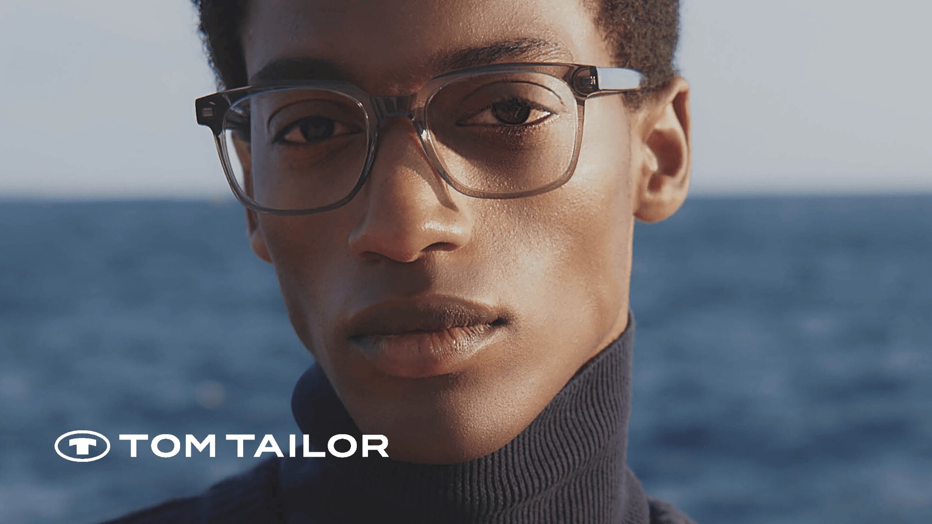 Eine Person mit einer Brille mit klarem Gestell und einem marineblauen Rollkragenpullover posiert im Freien mit dem Meer im Hintergrund. Das Tom Tailor-Logo erscheint in der unteren linken Ecke.