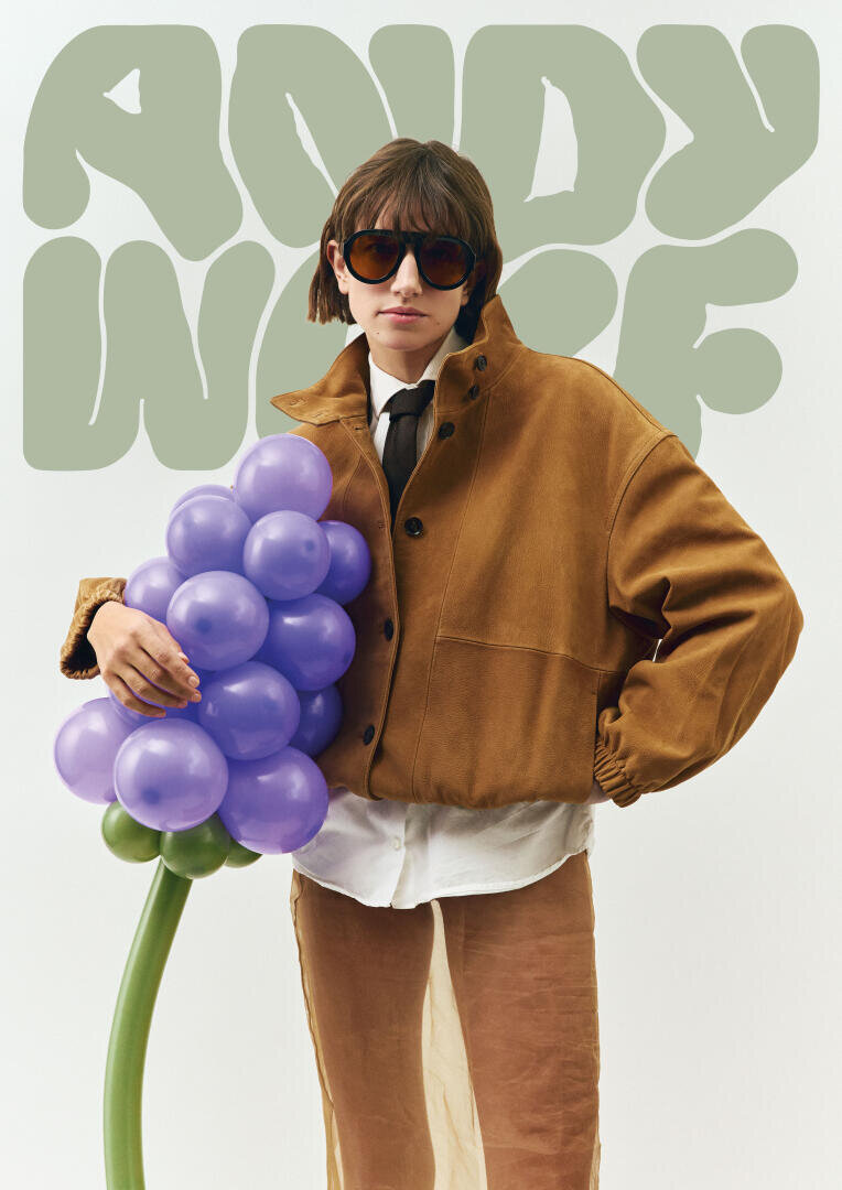 Eine Person mit Sonnenbrille, brauner Jacke und weißem Hemd hält einen Strauß lila Ballontrauben. Im Hintergrund ist ein abstrakter grüner Text mit der Aufschrift ANDY WOLF zu sehen.