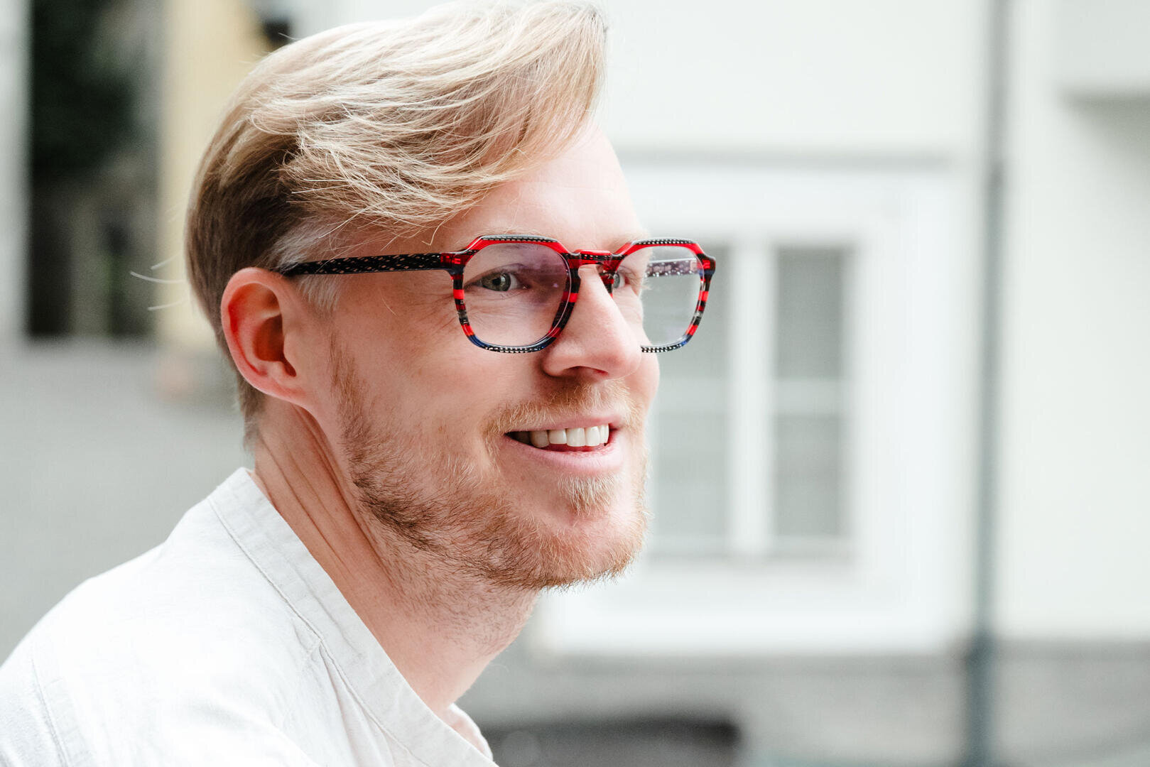Ein lächelnder Mann mit hellem Haar und Bart trägt eine auffällige rot-schwarz gemusterte Brille. Er trägt ein helles Hemd und steht im Freien vor einem verschwommenen Gebäude.