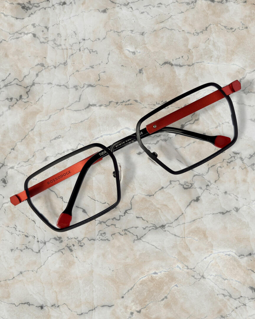 Eine rechteckige Brille mit schwarzem Rahmen und roten Bügeln ruht auf einer hellen Marmorfläche mit grauen und hellbraunen Äderungen.