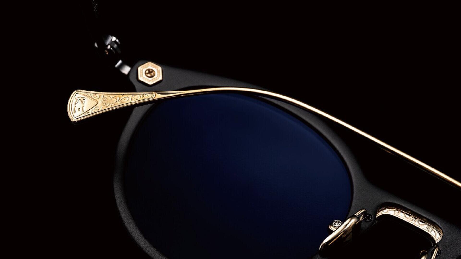 Close-up of a pair of luxury sunglasses, focusing on the gold-colored, intricately engraved temple and dark blue lens against a black background.