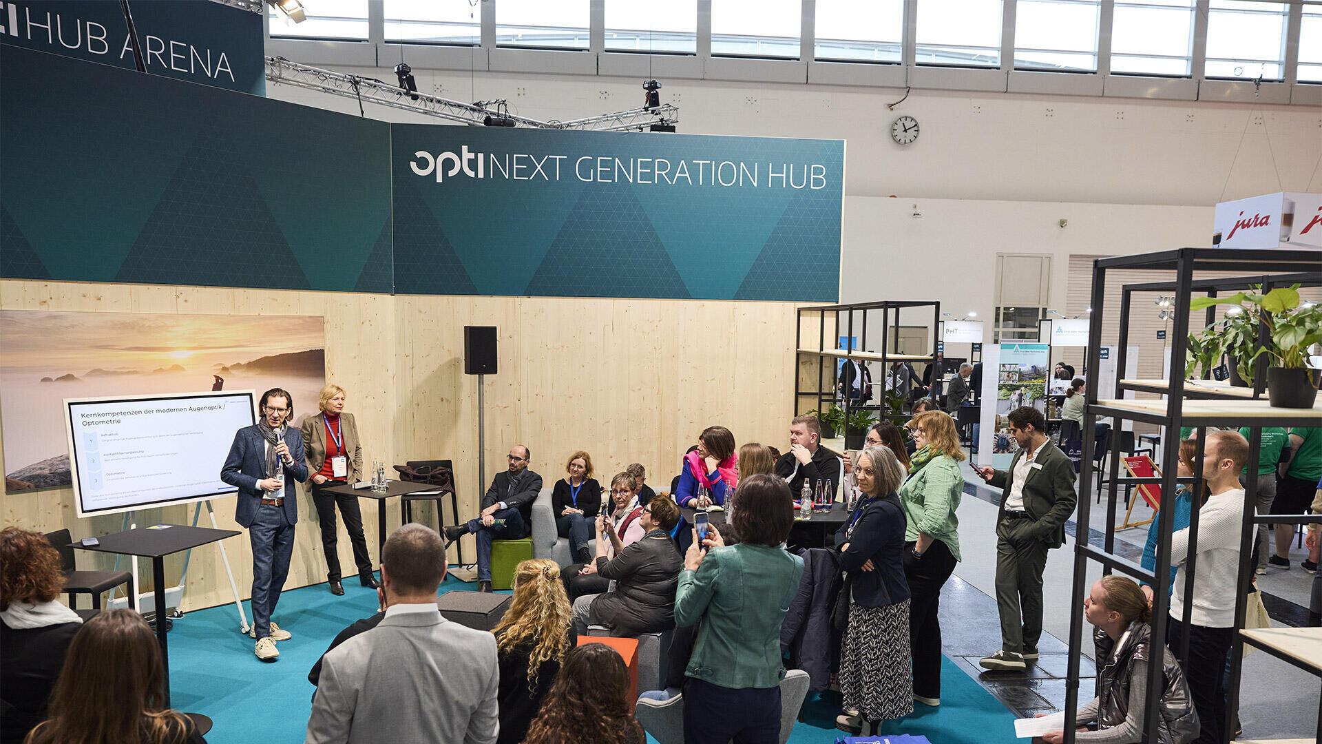 Ein Redner hält einen Vortrag vor einem aufmerksamen Publikum auf einer Konferenz in einem modernen, offenen Raum mit einem Bildschirm und einer Beschilderung mit der Aufschrift Next Generation Hub. Die Menschen sitzen und stehen, einige machen Notizen oder Fotos.